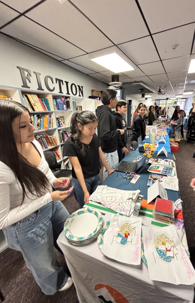 People gather around a table displaying books, plates, and a blue tablecloth. A woman looks at her phone.