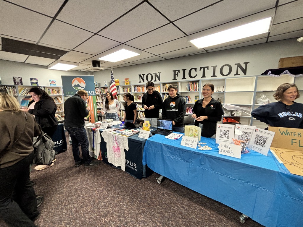 People stand around tables with blue cloths, displays, and signs. Behind them is a bookshelf with a sign that reads "Non Fiction".