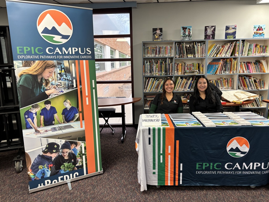 Two women sit at a table with pamphlets. A table has a blue cover with "EPIC CAMPUS" in white. Behind them is a bookshelf full of books.