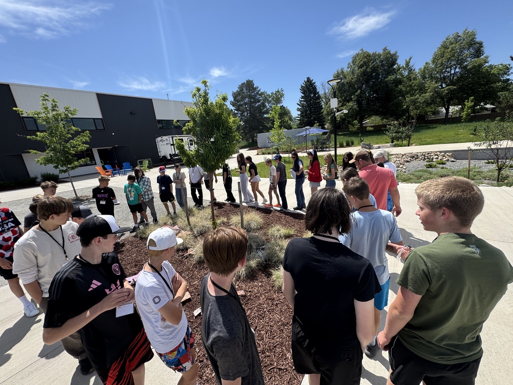 A group of people stand around a small tree in a landscaped area in front of a building.