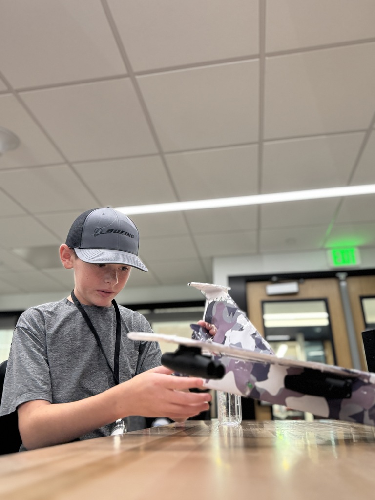 A young boy, wearing a hat and a gray shirt, works on a camouflage-patterned model airplane at a table.