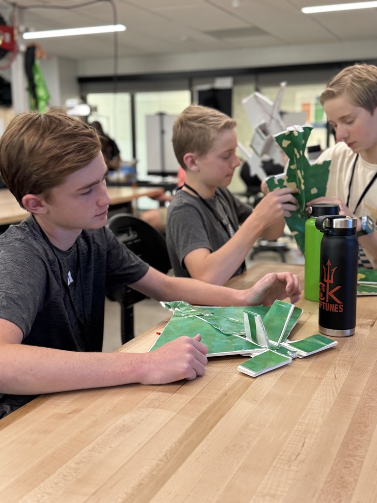Three boys work on a project at a table in a room with white walls and ceiling lights.
