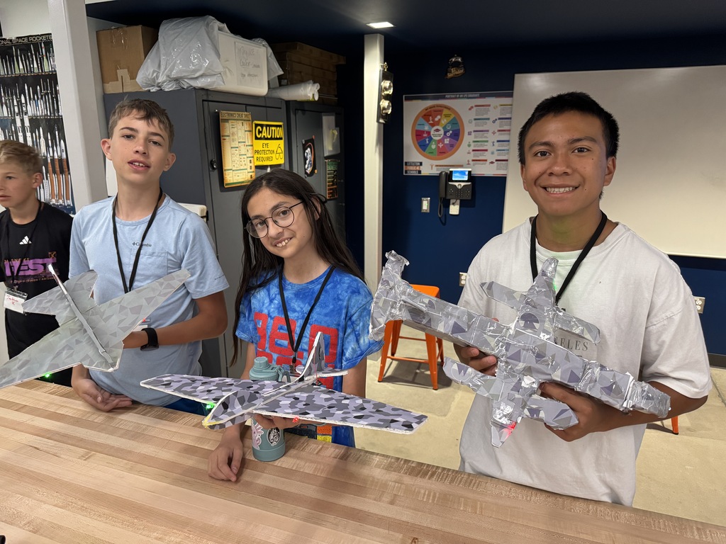 Three young people posing with airplane models in a classroom, one wearing glasses, another with a blue shirt.