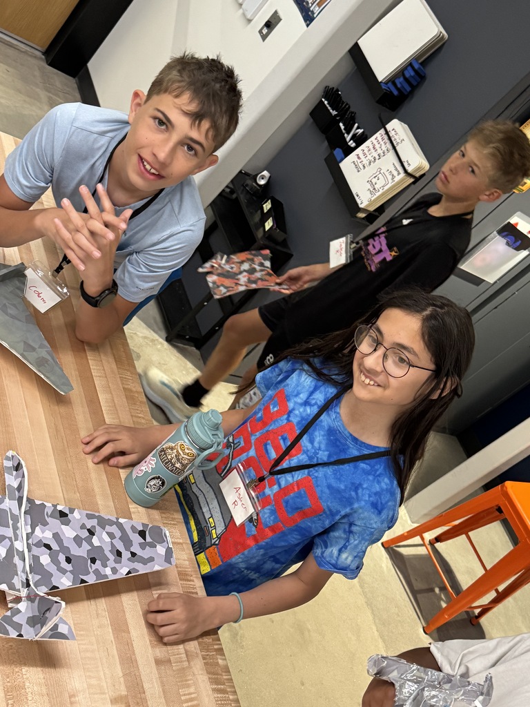 Three kids at a table, one smiling, another holding a book, and the third with glasses. Background includes shelves with books and an orange chair.