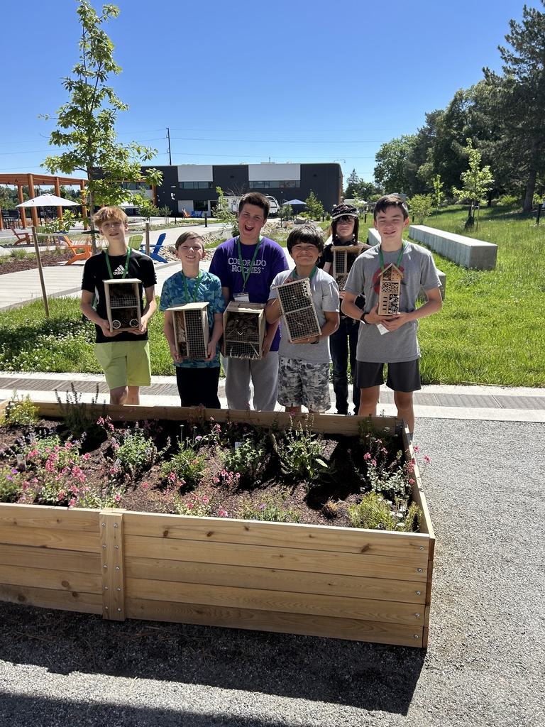 Seven kids stand in a raised garden with wooden planters, holding small wooden boxes. A building is in the background.