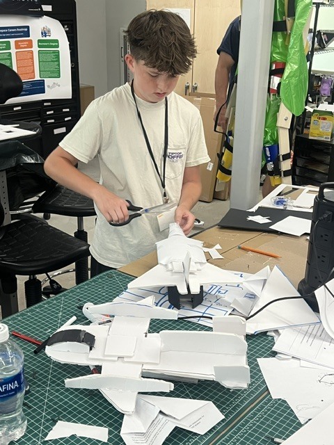 A boy works on a project at a table, surrounded by scattered paper and tools, with a person partially visible in the background.