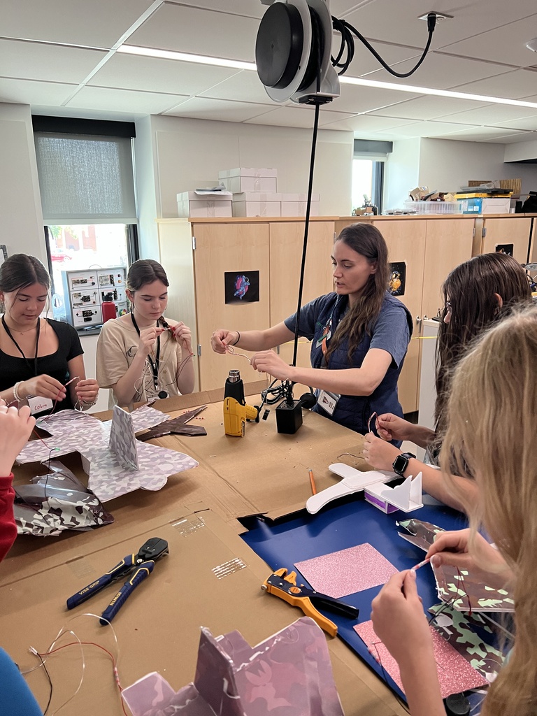 A group of people work on a project in a classroom. A woman stands at the front with a blue table covered in materials.
