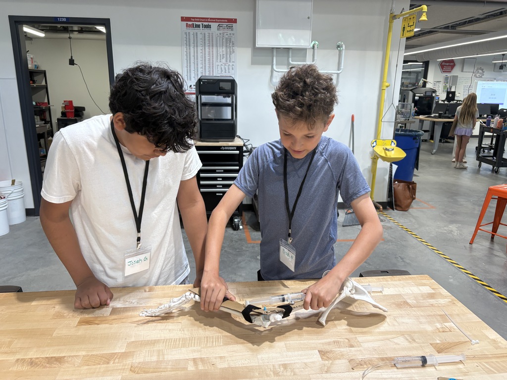 Two children in lab coats examine a piece of equipment on a wooden table in a workshop.