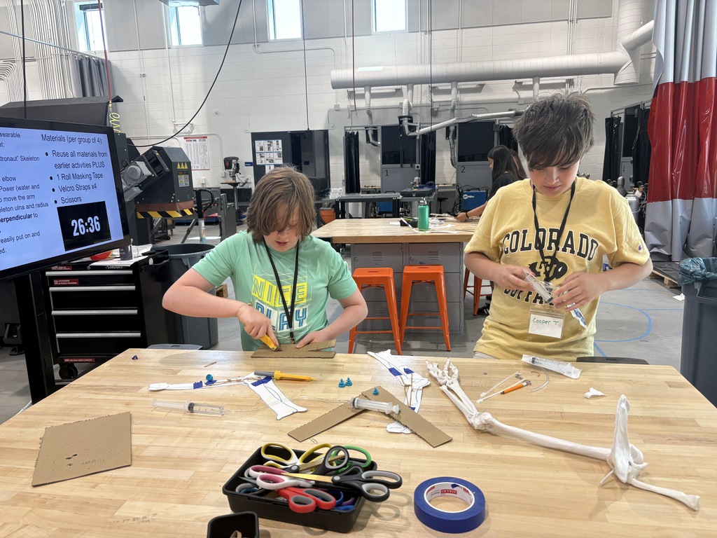 Two children in a workshop use tools to work on a project at a wooden table.