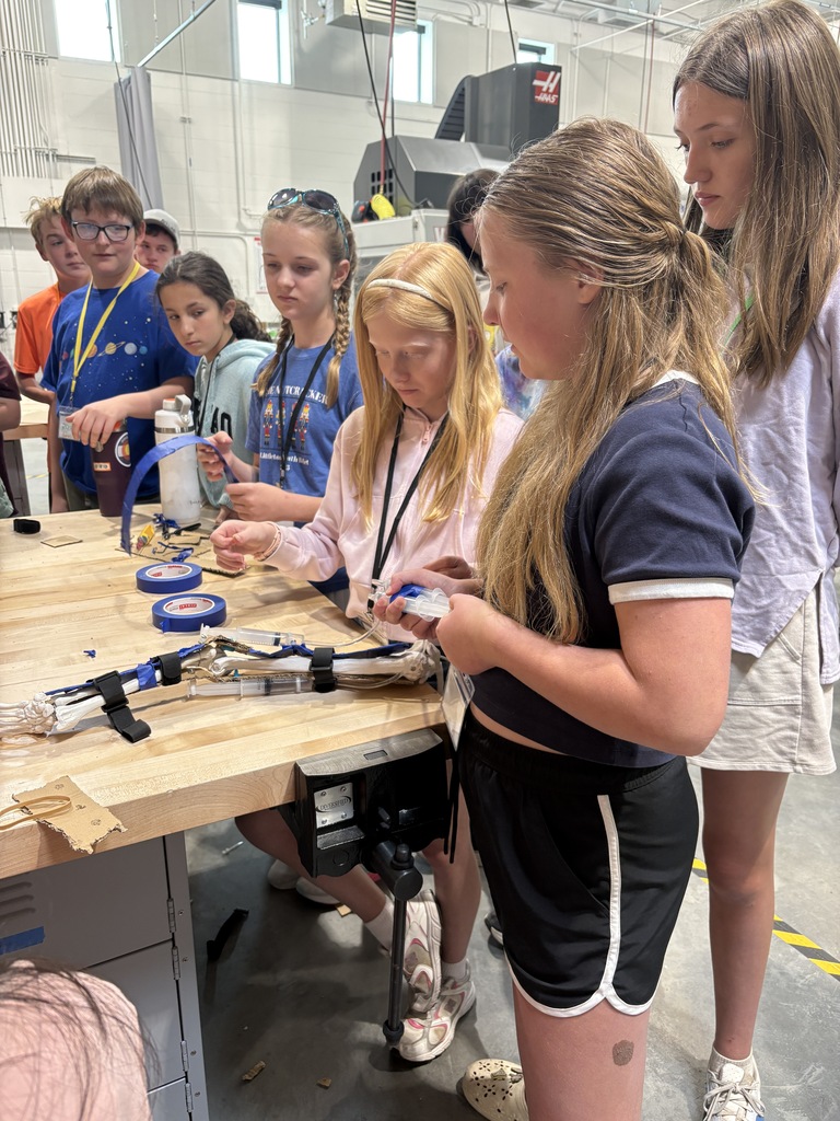Several young girls work on a project at a table in a workshop; tools and materials are visible.