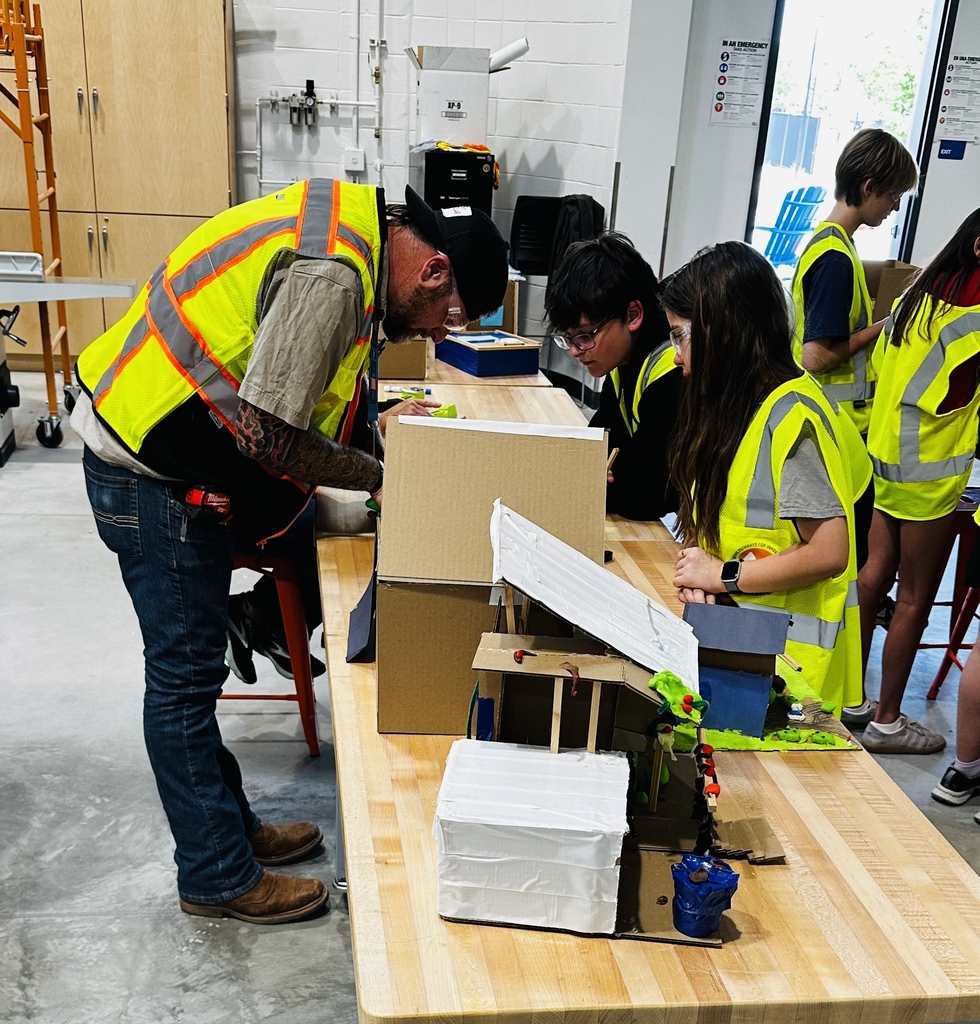 A group of people in safety vests working on a project. A man works on a box while others look on.