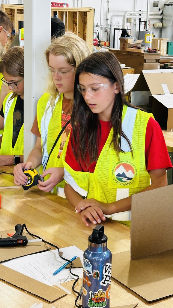 Three girls in yellow vests work on a wooden desk with tools, papers, and a bottle in a workshop.