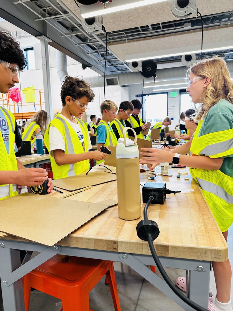A group of kids in yellow vests work on a project at a table in a workshop.