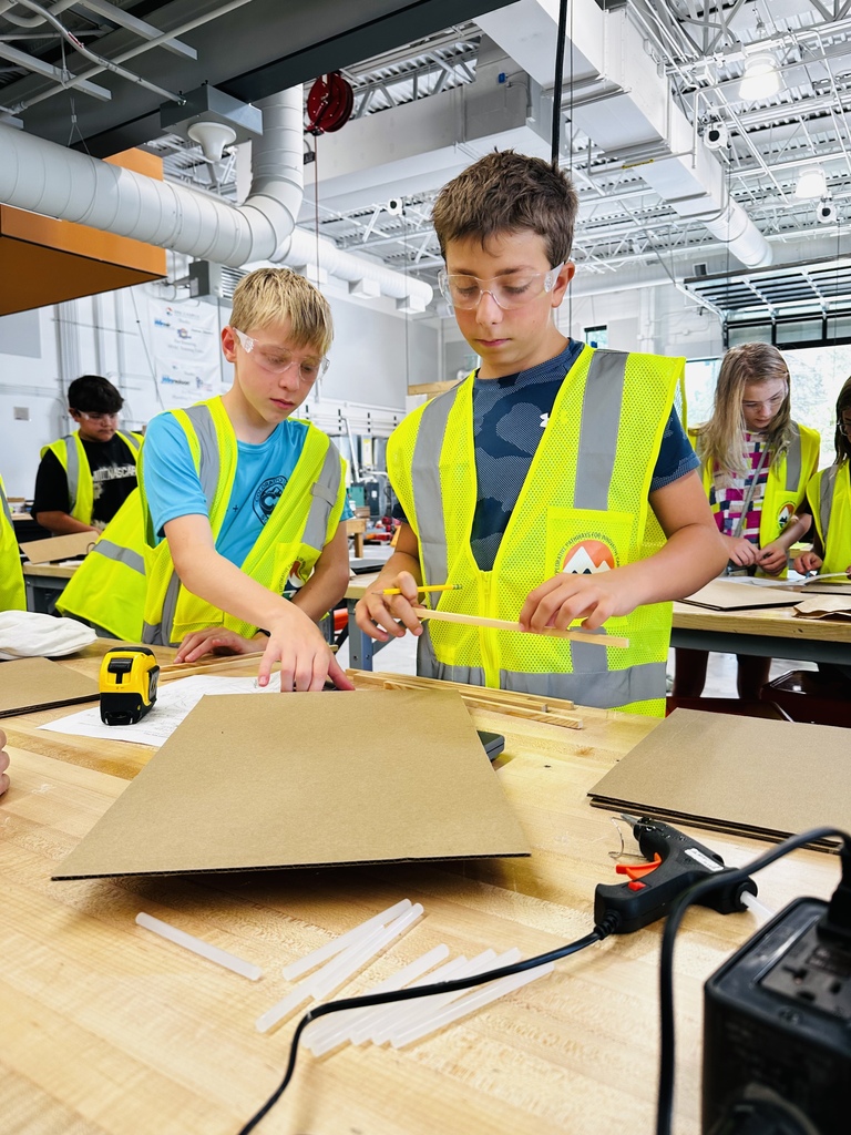Kids wearing yellow vests work on a project at a workbench in a room with bright lights.