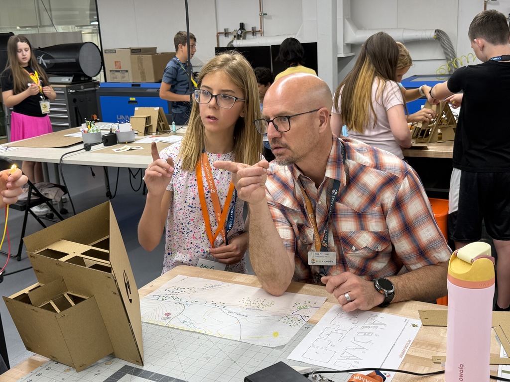 A man and a girl are examining a blueprint on a table; other people are working in the background.