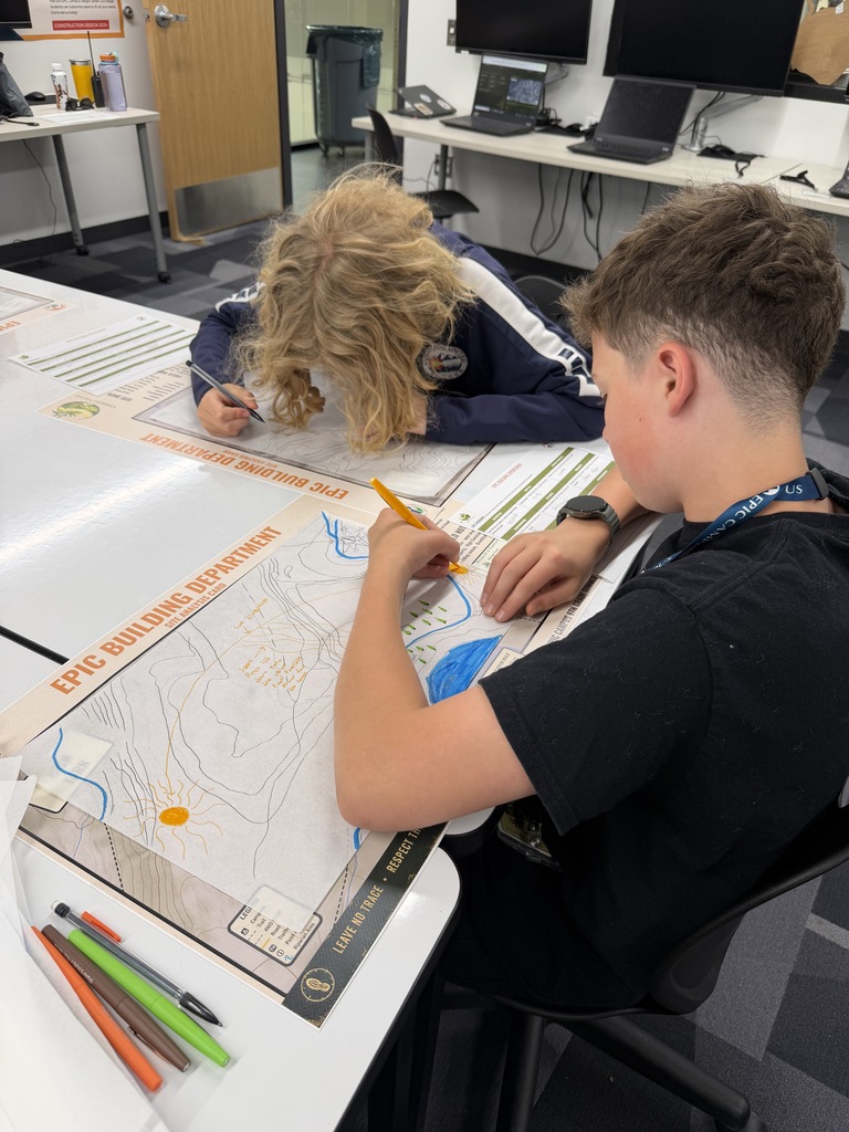 Two students working on a large map, sitting at desks with computers, pens, and pencils.