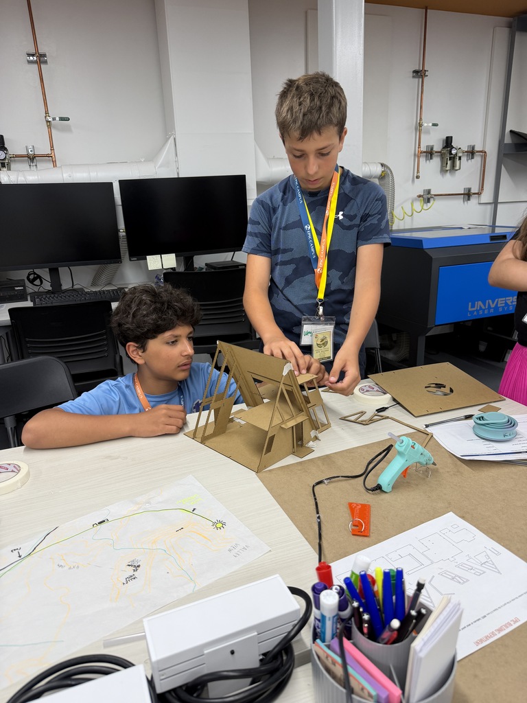 Two young boys working on a cardboard structure on a table in a classroom with computers.