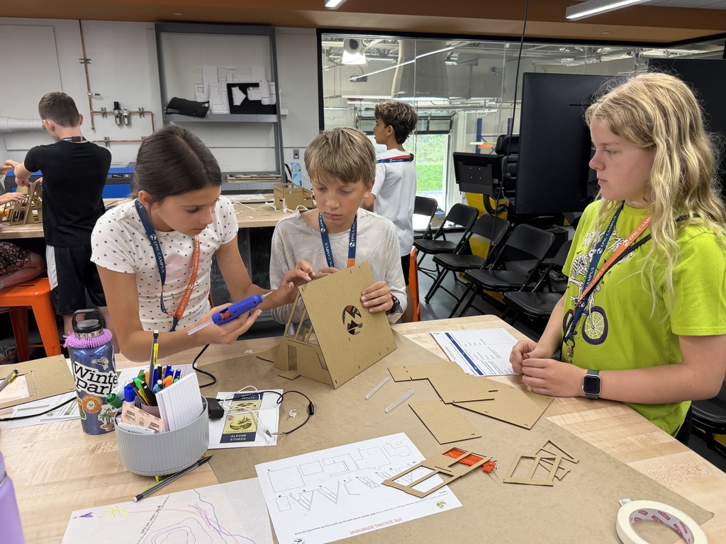 Three children in a room with white walls, working on a cardboard project on a table.