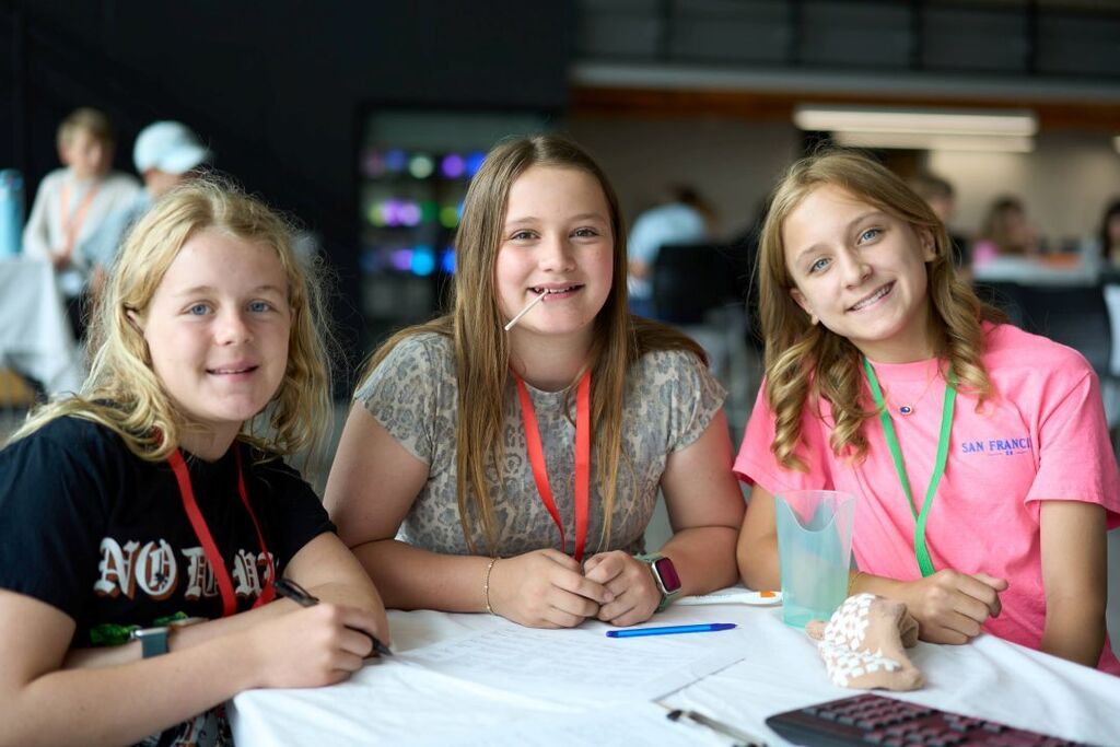 Three girls sit at a table, smiling, with two holding pens and one holding a shell.