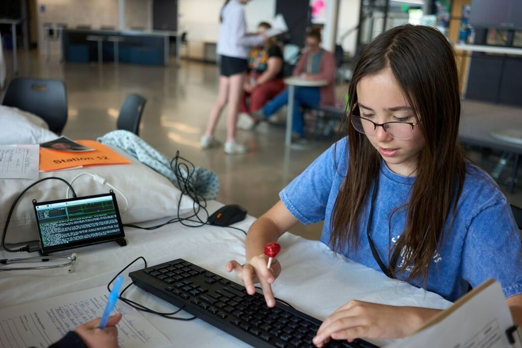 A girl with glasses seated at a table, typing on a keyboard while looking at a screen.