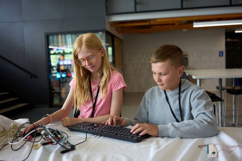 Two kids seated at a table with a keyboard and wires, working on a computer project.