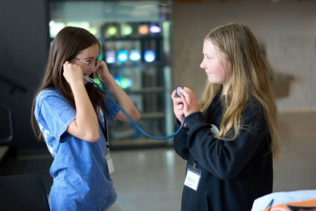 Two girls in a room. One girl has a stethoscope around her neck, and the other has a stethoscope on her ear.