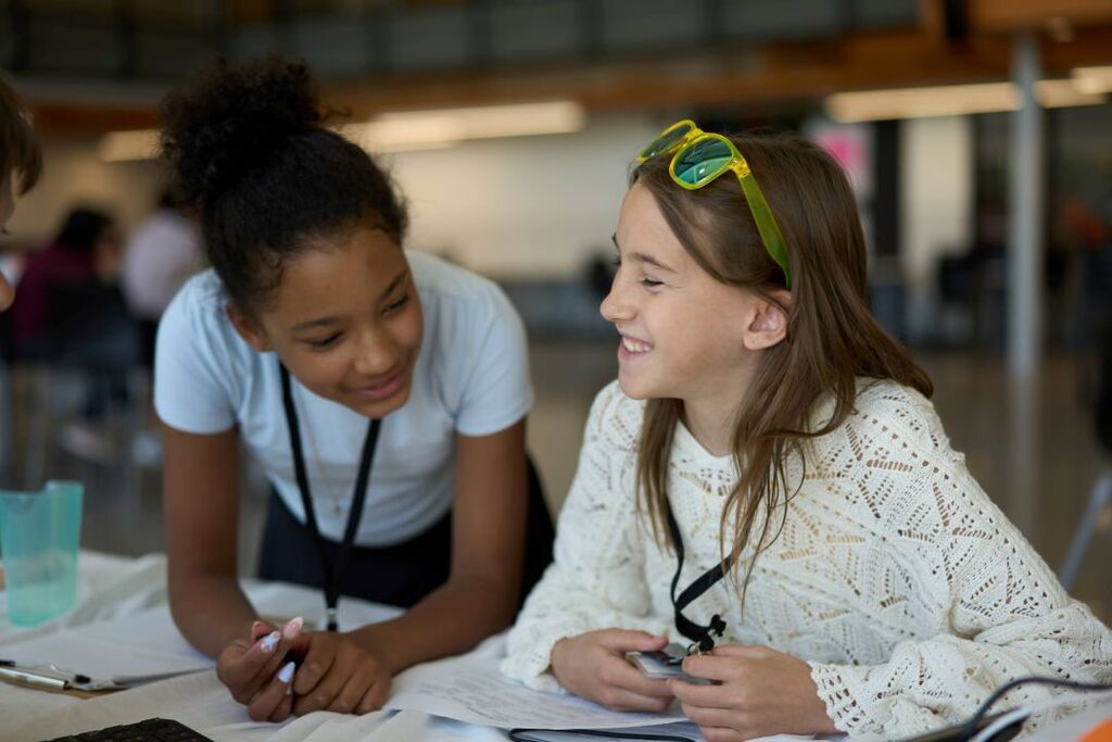 Two girls sit at a table with papers and pens, smiling and talking to each other. They are in a room with fluorescent lights.