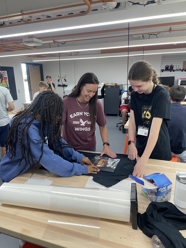Three people work on a table with a rolled up material, a black t-shirt, and other items.