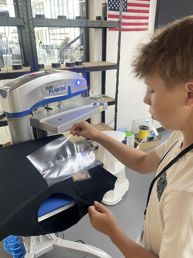 A child in a light shirt places a shirt on a machine in a factory. An American flag hangs on the wall.