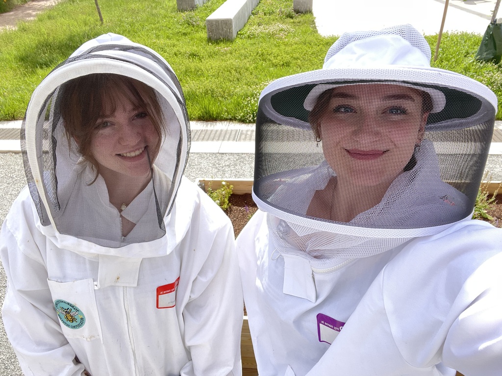 Two women in beekeeping suits and hats standing together outside with a grassy background.