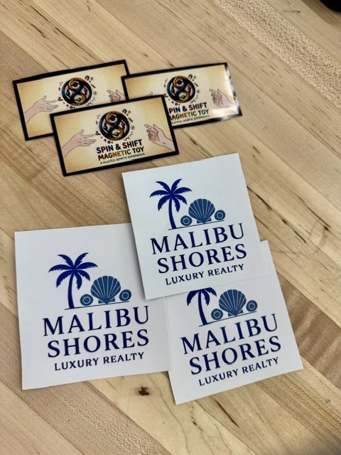 Four rectangular labels on a wooden surface. Each label has "Malibu Shores Luxury Realty" written in blue. Two labels feature palm trees and seashells.