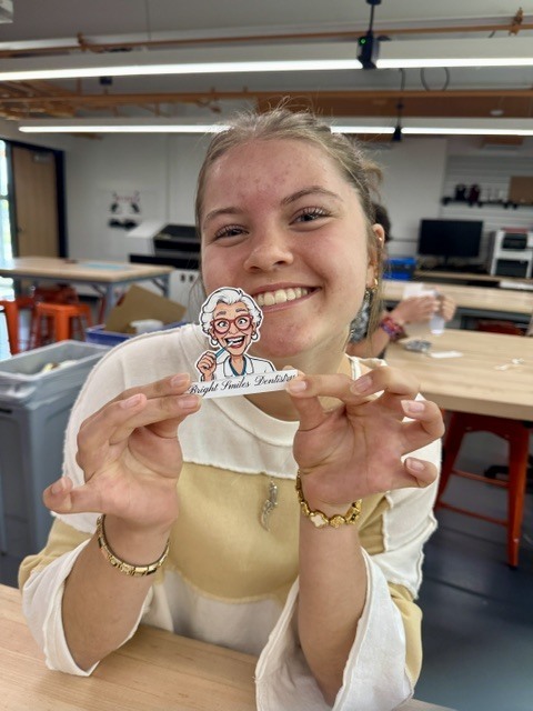 A woman holds a sticker of an elderly woman, smiling and seated at a table in a workshop.