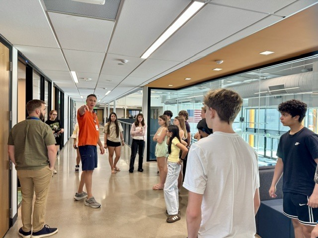 Several people stand in a hallway. A man in an orange shirt faces away from the group. A large window is behind them.