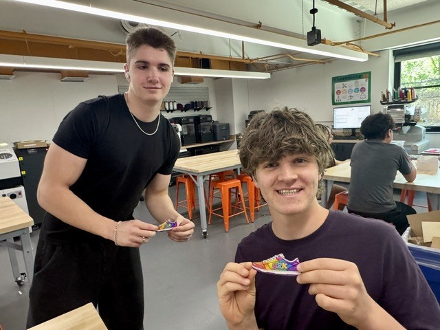 Two young men in a workshop. One holds a shoe and the other holds a shoe. Behind them, other people work at tables.
