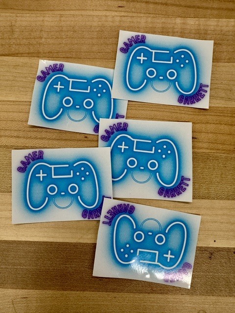 Four stickers with blue game controller designs are displayed on a wooden surface. Each sticker features a unique name below the controller.