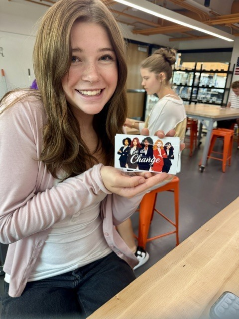 A person smiles while holding a card with images of a group of women and text.