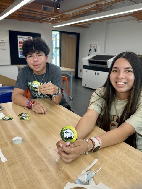 Two young people seated at a wooden table. One holds a yellow button. Behind them is a printer and a white door.