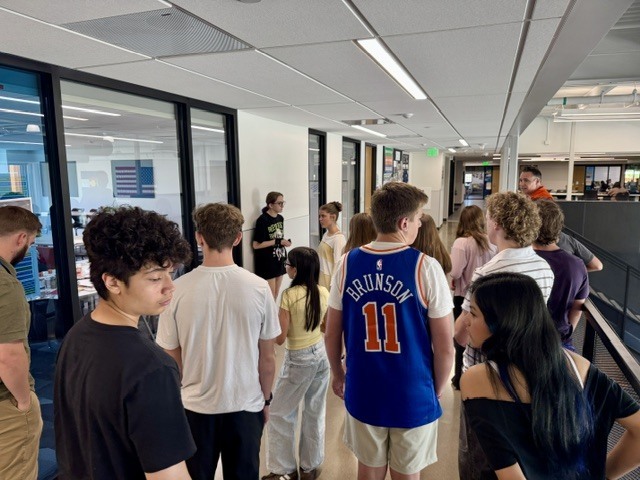 Several people stand in a hallway, some wearing face masks. An American flag is visible through a window.