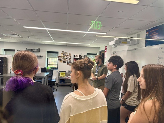 A classroom with bright lights, where people in casual clothing are gathered. A man stands in front, addressing the group.