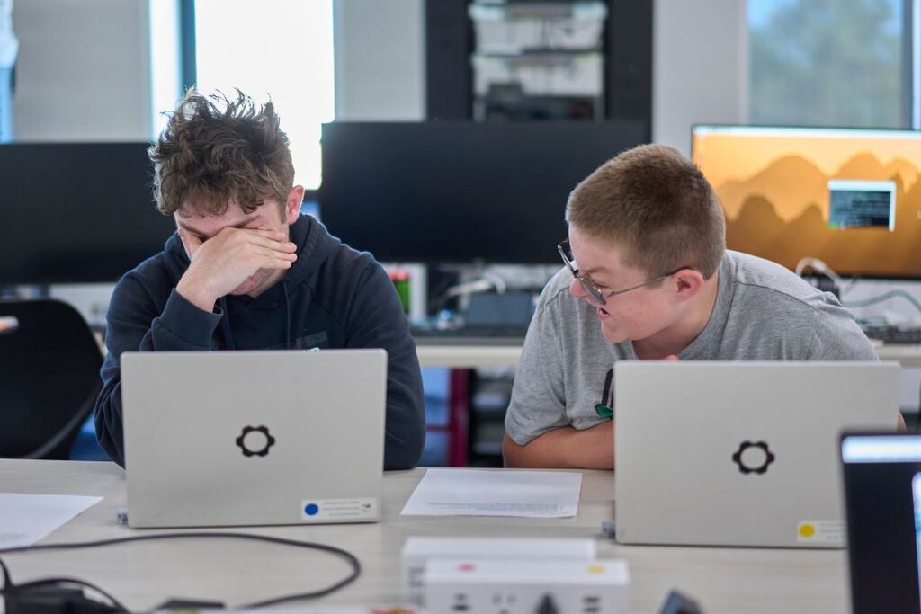 Two people sit at a table with laptops; one wearing glasses and the other with hand on face.