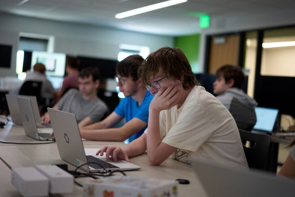 Four students seated at a desk, using laptops. One student wears glasses and rests their hand on their face.