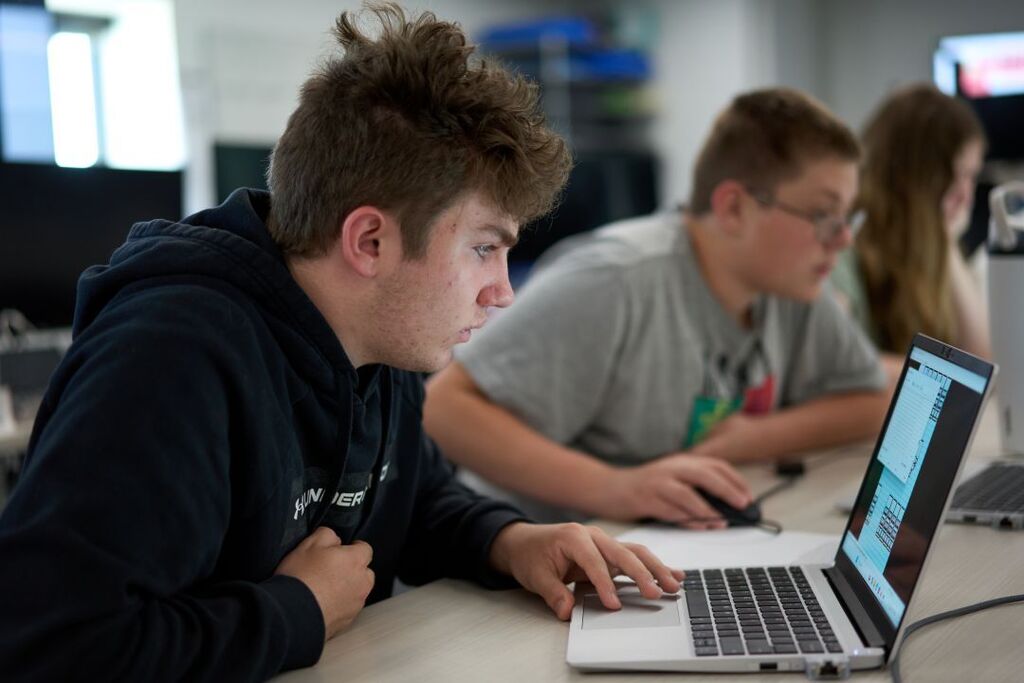 Three young people work on laptops in a classroom; one wears a black hoodie, one a gray shirt.