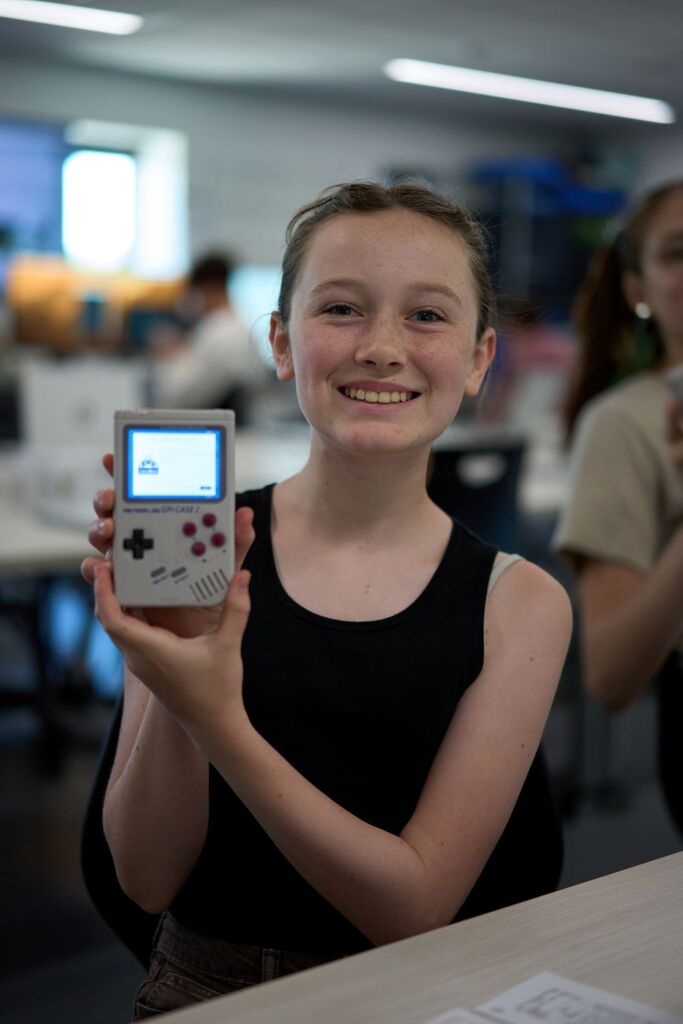 A girl in a black tank top holds a small handheld video game device, smiling in a classroom.