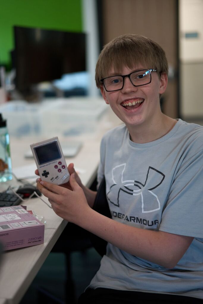 A boy wearing glasses and a gray Under Armour shirt holds a Gameboy Advance in a classroom.