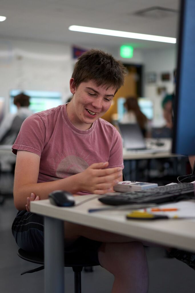 A person in a pink shirt sitting at a desk using a computer. Desk has a keyboard, mouse, and papers.