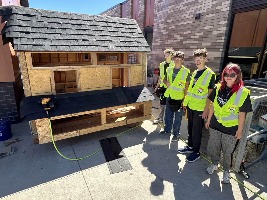 Four people in reflective vests stand next to a wooden house-like structure. The house has a roof and a black lining.