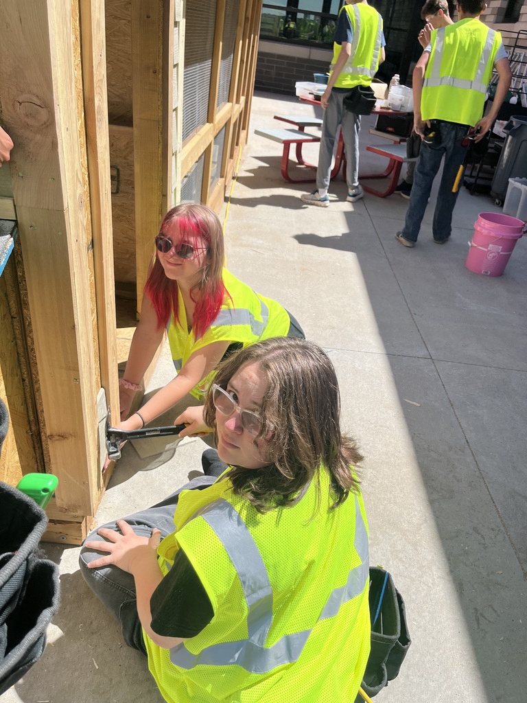 Two people in yellow vests work on a wooden box. Other people are in the background wearing vests.