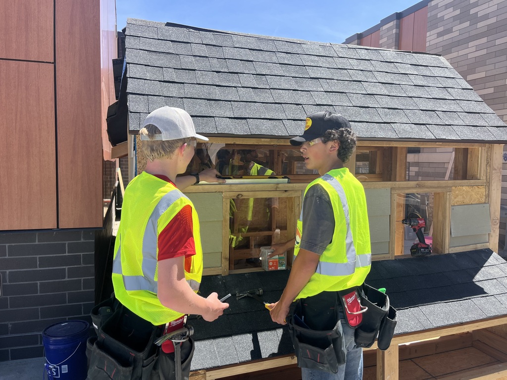 Two people in reflective vests and hats are standing in front of a model house. One looks at a phone.
