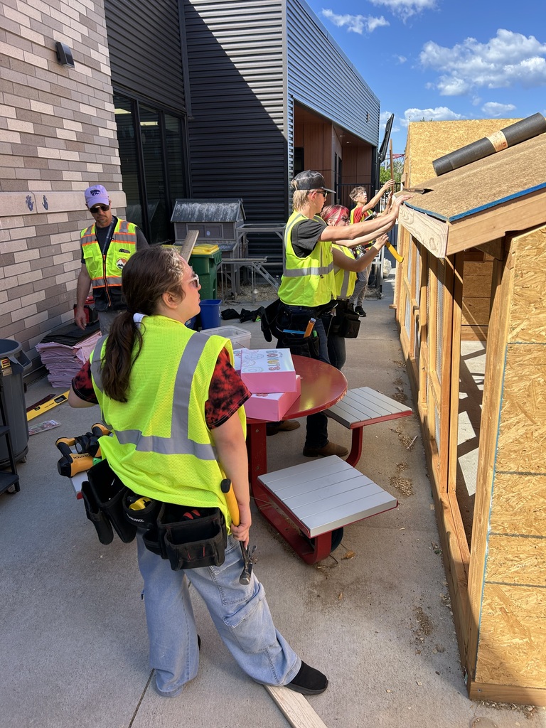 A group of people in yellow vests work on a wooden structure outdoors. A building is visible in the background.