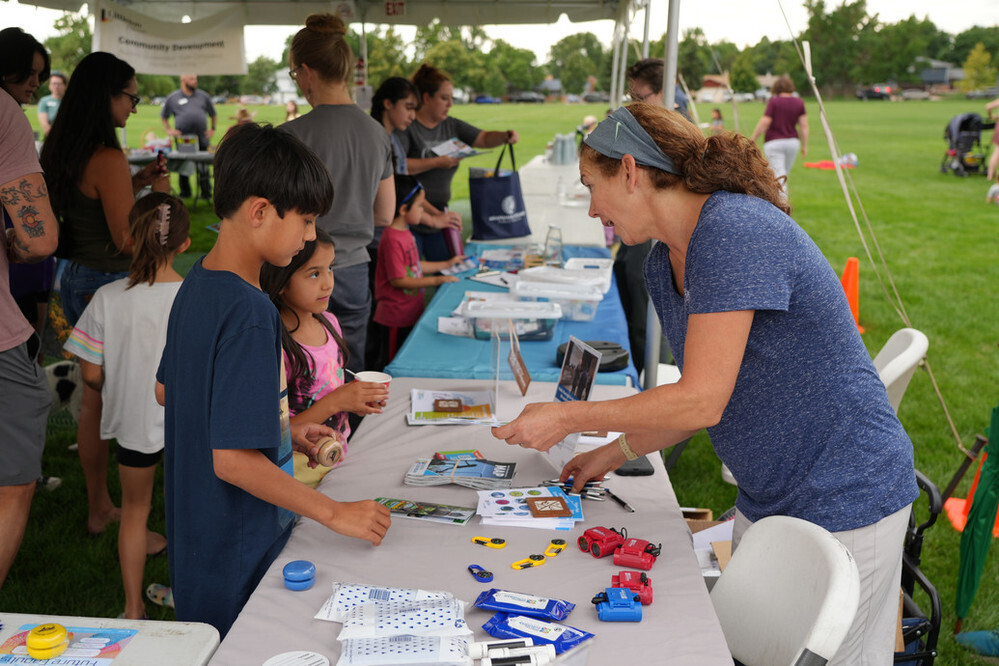 A woman hands out materials to a child at an event on a grassy field. Other adults and children stand nearby.